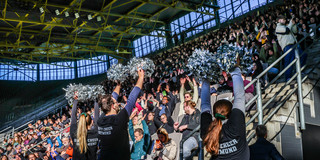 Auf dem Foto sieht man, wie das Publikum und die Cheerleader*innen gemeinsam eine La-Ola-Welle machen.