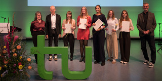 Foto: Acht Personen stehen auf einer grün beleuchteten Bühne vor einer Leinwand mit Titeltext, fünf von ihnen halten Papiere und Blumen, im Vordergrund steht ein grünes „tu"-Logo.