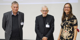 Auf dem Foto stehen drei Personen bei einer Veranstaltung nebeneinander und lächeln in die Kamera. Die beiden Männer (links) sind in Anzügen gekleidet, während die Frau (rechts) ein schwarz-rotes Kleid trägt.