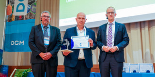 DGM-Preis Prof. Walther Drei Männer vor einer Präsentation. Der Preisträger Prof. Walter steht in der Mitte und hält Pokal und Urkunde in den Händen.