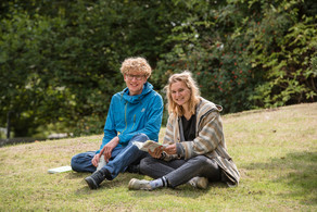 Campus Ein blonder Student mit blauer Jacke und eine blonde Studentin mit brauner Jacke sitzen auf einer Wiese