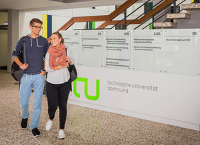 Studieninteressierte Ein Studentenpärchen läuft an einem TU-Logo im Foyer eines Gebäudes vorbei.